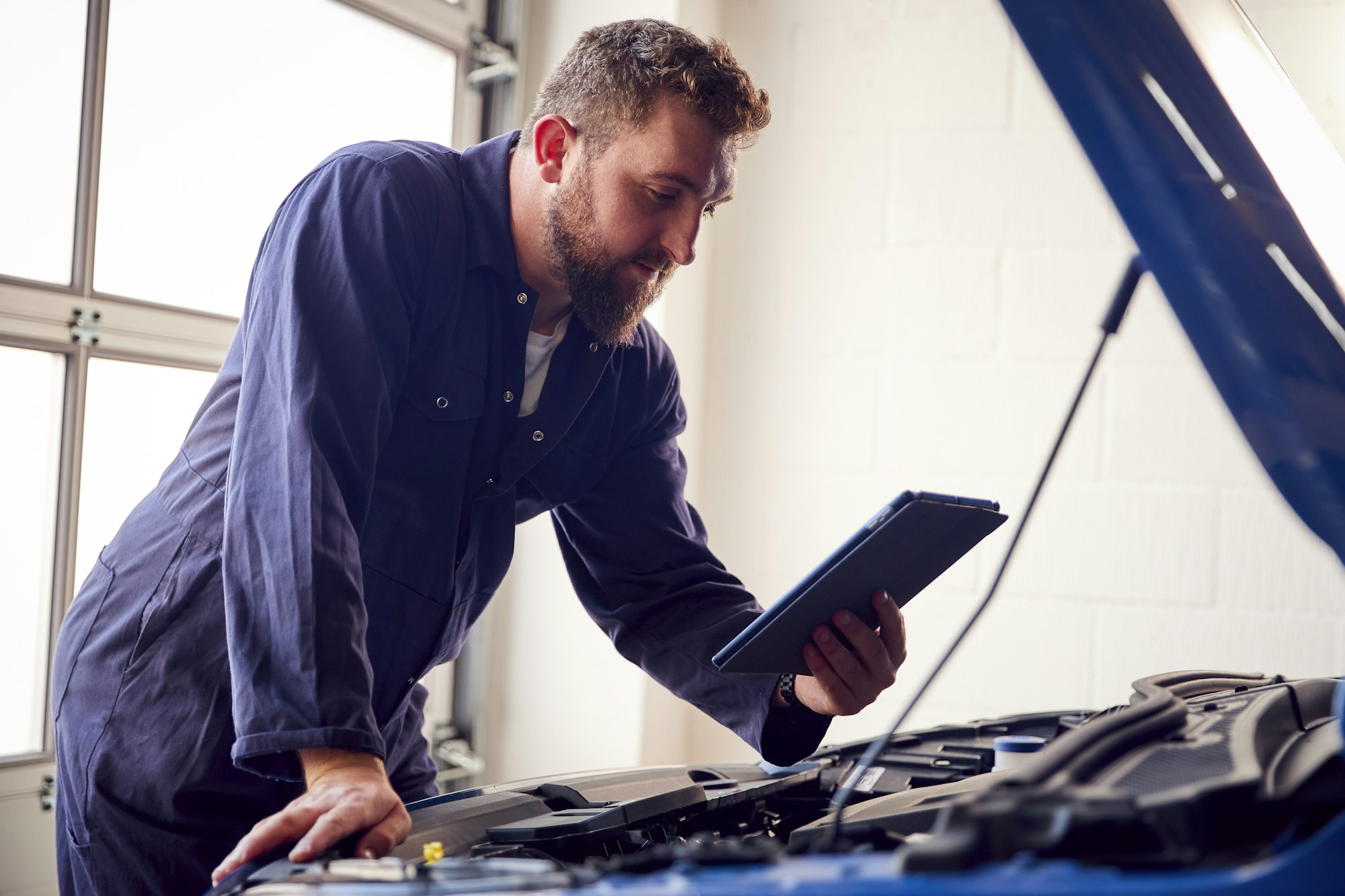 Male Mechanic In Garage Using Diagnostic Software On Digital Tablet To Service Car Engine